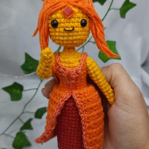 Op de afbeelding: Handgemaakte gehaakte pop met een personage met oranje garen haar en een bijpassende jurk. De pop heeft een geel gezicht met zwarte ogen en een kleine glimlach. De jurk is oranje en rood, met een ruitvormig accent.
