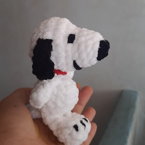 Può includere: Peluche di cane bianco all'uncinetto con orecchie e naso neri. Il cane indossa un collare rosso e ha un'espressione felice.