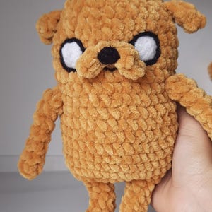 Puede incluir: Un juguete de peluche de crochet de un perro marrón con ojos blancos y una nariz negra. El juguete está de pie sobre una superficie blanca.