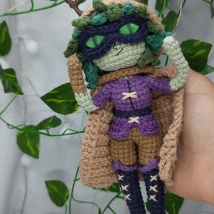 Puede incluir: Muñeca de ganchillo hecha a mano con un diseño caprichoso. La muñeca presenta una cara verde, un atuendo morado y una capa marrón. Tiene una máscara y ojos verdes. La muñeca está adornada con detalles verdes y marrones, con temática de fantasía.