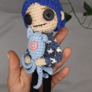 May include: Crocheted doll with blue hair, button eyes, and a stitched mouth. The doll wears a blue sweater with white star accents, black trousers, and blue shoes. It holds a blue stuffed animal with a pink button.