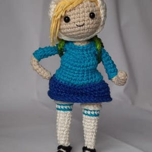 Pode incluir: Boneca de crochê de um personagem de desenho animado com um chapéu branco com orelhas, cabelo loiro e um vestido azul. A boneca tem sapatos pretos e meias brancas com detalhes azuis. A boneca tem cerca de 20 cm de altura.