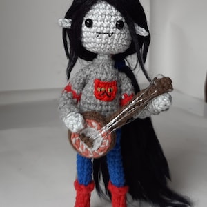 Puede incluir: Una muñeca de crochet con cabello negro largo, vistiendo un suéter gris con un diseño de gato rojo, pantalones azules y botas rojas. La muñeca sostiene un banjo marrón.