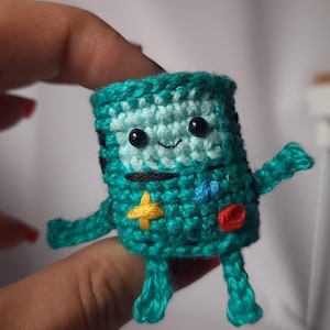 Op de afbeelding: Een klein, gehaakt groen figuurtje met een glimlachend gezicht, zwarte ogen en gele en rode details. Het figuurtje heeft vier ledematen en doet denken aan een cartoonfiguur.