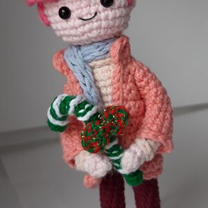 Pode incluir: Boneca de crochê feita à mão com casaco, chapéu e cabelo rosa, cachecol azul e calças bordô. A boneca está segurando um bastão de doces verde e branco e um bastão de doces vermelho e verde. A boneca está usando sapatos azuis e brancos.