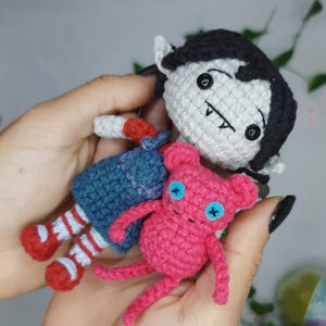May include: Crocheted dolls of a vampire character with black hair and a pink creature with blue cross-shaped eyes. The vampire doll wears a blue dress and red and white striped socks. The pink creature has small arms and legs.