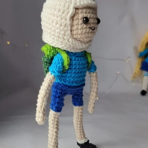 May include: Crocheted Finn the Human from Adventure Time, wearing a white hat with ears, a blue shirt, blue shorts, and a green backpack.