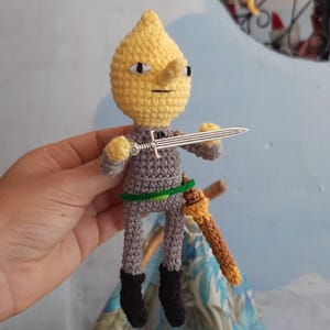 Puede incluir: Un juguete de peluche tejido a crochet de un personaje amarillo con un atuendo gris, sosteniendo una espada plateada y una espada marrón. El personaje tiene una nariz puntiaguda y una expresión seria.