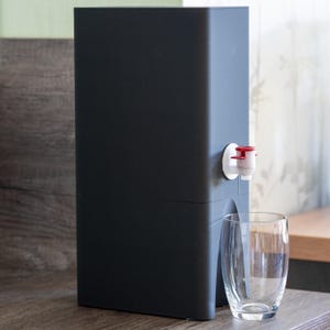 Könnte beinhalten: Ein dunkelgrauer Wasserspender mit einem weißen und roten Hahn. Ein klares Glas steht vor dem Spender.