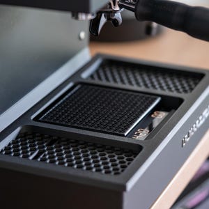 Op de afbeelding: Close-up van een zwarte espressomachine met een ingebouwde weegschaal en manometer. De machine heeft een portafilter en een lekbak met een honingraatpatroon. De weegschaal geeft een digitale aflezing.