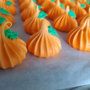 May include: Rows of orange-colored meringue cookies shaped like pumpkins, each with a green leaf detail on top. The cookies are arranged on a white surface, creating a festive and seasonal treat.