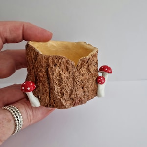 May include: A small tree stump planter with a textured brown exterior and a light yellow interior. Two small mushrooms with red caps and white spots are attached to the side. Ideal for miniature plants or fairy gardens, this item has a whimsical design.