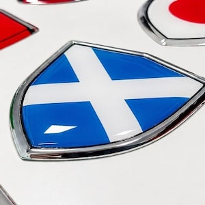 Op de afbeelding: Een verzameling schildvormige emblemen met diverse nationale vlaggen, voorzien van een glanzende, bolle afwerking en een zilverkleurige metalen rand. Het centrale embleem toont de blauw-witte vlag van Schotland, omringd door andere ontwerpen.