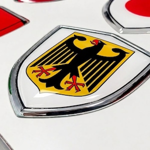 Peut inclure: Un emblème bombé en forme de bouclier représentant l'aigle allemand noir sur fond jaune, bordé d'un champ blanc et d'un cadre en argent métallique. Le design est brillant et en relief, placé parmi d'autres emblèmes de drapeaux nationaux.