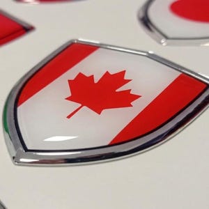 Op de afbeelding: Schildvormige emblemen met nationale vlaggen. Het Canadese vlagembleem staat centraal, met een rood esdoornblad op een witte achtergrond met rode strepen. Andere vlaggen zijn China en Japan. Elk embleem heeft een zilveren rand.