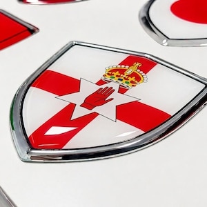 Könnte beinhalten: Ein schildförmiges Emblem mit der Flagge von Nordirland, bestehend aus einem roten Kreuz, einem weißen sechsstrahligen Stern, einer roten Hand und einer Krone. Das Emblem hat eine glänzende Oberfläche und einen metallisch silbernen Rand.