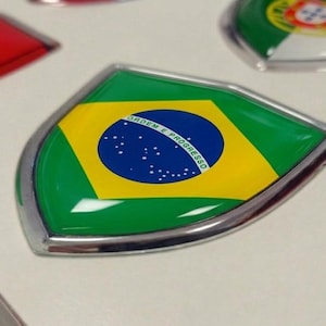 Puede incluir: Un emblema en forma de escudo con la bandera verde, amarilla y azul de Brasil, acabado brillante y borde metálico plateado. El círculo azul central contiene estrellas blancas y el texto ORDEM E PROGRESSO en letras mayúsculas de color blanco.