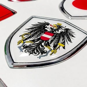 Op de afbeelding: Een schildvormig embleem met het Oostenrijkse wapen, voorzien van een zwarte adelaar, een rood-wit-rood borstschild en gouden klauwen die een hamer en sikkel vasthouden, gevat in een glanzende, zilverkleurige metalen rand.