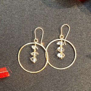 Puede incluir: Un par de pendientes de aro dorados con detalles de cristal transparente que cuelgan de la parte inferior. Los pendientes están sobre un fondo de tela negro.