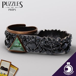 May include: A black and green ring with a triangular emerald gemstone set in a bed of black stones. The ring is made of metal and has a textured surface.