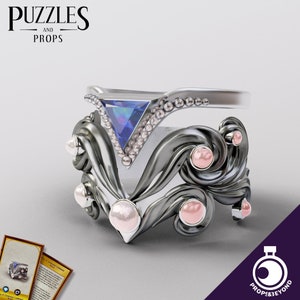 May include: A silver ring with a triangular blue gemstone and small clear stones. The ring has a dark gray, ornate base with pink and white pearl-like accents. The text "PUZZLES AND PROPS" is at the top.