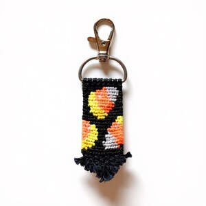 May include: A rectangular macrame keychain featuring a black background with a repeating pattern of candy corn shapes in yellow, orange, and white. The woven textile is attached to a silver-colored metal lobster clasp and a D-ring, with a frayed fringe.