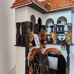 May include: A miniature wooden house with a white exterior, brown trim, and a red tile roof. The house has two balconies with black railings and potted plants. The front door is black with a gold-colored design. The house has a sign that says "Colombia" on the side.