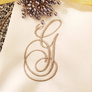 May include: A white linen napkin with a large embroidered letter G in a script font. The letter is in a light brown color. A decorative cluster of gray pearl beads is on top of the napkin.