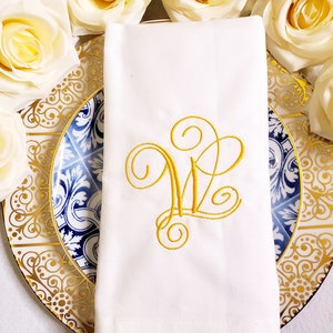 Puede incluir: Servilleta de lino blanco con un monograma bordado en oro de la letra "W" sobre un fondo blanco. La servilleta está doblada y colocada sobre un plato con estampado azul y blanco con ribete dorado.