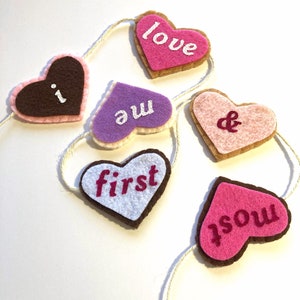 Puede incluir: Una guirnalda de fieltro con seis corazones, cada uno con una palabra diferente. Las palabras son "i", "love", "you", "&", "first" y "most". Los corazones son de varios colores, incluyendo rosa, marrón, morado y blanco.