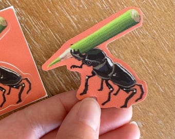 Peut inclure: Un autocollant représentant un scarabée noir tenant un crayon vert. Le scarabée est sur un fond orange. L'autocollant est tenu par une main.