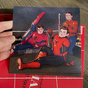 May include: Three cartoon illustrations of Spider-Man in red and blue suits with black spider emblems. The figures are posed in a relaxed manner on a rooftop with a night sky background.