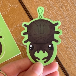 May include: A green and black beetle-shaped sticker with a loop at the top. The beetle has a dark brown body and large, cartoonish eyes. The sticker is held by a hand, with a wooden surface in the background.