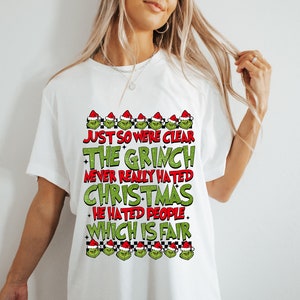 May include: White t-shirt with a Grinch-themed design. The text reads "JUST SO WE'RE CLEAR THE GRINCH NEVER REALLY HATED CHRISTMAS HE HATED PEOPLE WHICH IS FAIR" in green and red. The design includes Grinch faces wearing Santa hats.
