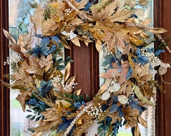 Puede incluir: Una corona decorativa con una mezcla de hojas artificiales doradas, azules y crema. La corona incluye varias formas y texturas de hojas, con pequeños acentos de bayas blancas. Se exhibe en una puerta de madera marrón con paneles de vidrio.