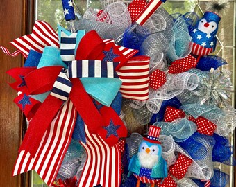 Puede incluir: Una corona patriótica con cintas de malla rojas, blancas y azules, un gran lazo con patrones de estrellas y rayas, dos adornos en forma de fuegos artificiales y dos pequeñas figuras de pájaros con sombreros de copa en una disposición circular.