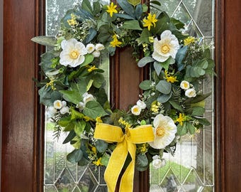 Puede incluir: Una corona de primavera con flores blancas y amarillas, hojas verdes y una cinta amarilla. La corona está colgada en una puerta marrón con paneles de vidrio.