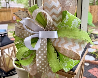 Puede incluir: Un lazo decorativo hecho de varias cintas con patrones florales verde lima, estampados de chevrón en blanco y tostado, lunares tostados y una cinta de satén blanca sólida. El diseño en capas incluye bordes con alambre y texturas de arpillera.