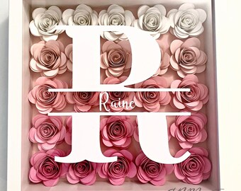 May include: A white shadow box with a white letter R and the name "Raine" centered in the middle. The background is filled with pink and white paper flowers.