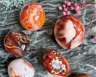 Puede incluir: Siete esferas de cristal de cornalina de diferentes tamaños y patrones, con colores naranja, rojo y blanco, dispuestas sobre una superficie de mármol verde y blanco.