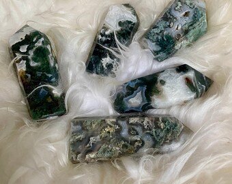 May include: Five green, white and grey moss agate crystal points. The points are arranged in a cluster on a white fluffy surface.