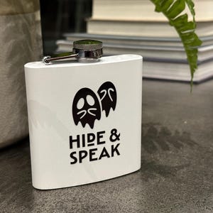Könnte beinhalten: Weißer Flachmann mit schwarzer Geistergrafik und den Worten "HIDE & SPEAK". Der Flachmann hat einen silbernen Verschluss und ein Scharnier. Der Flachmann steht auf einer dunklen Oberfläche.