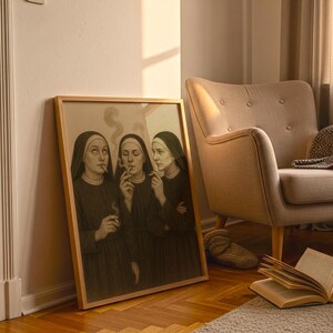 Puede incluir: Una obra de arte enmarcada representa a tres monjas fumando cigarrillos. Las monjas visten h&aacute;bitos negros y tocas blancas. La obra est&aacute; apoyada contra una pared junto a un sill&oacute;n beige y una mesita auxiliar.