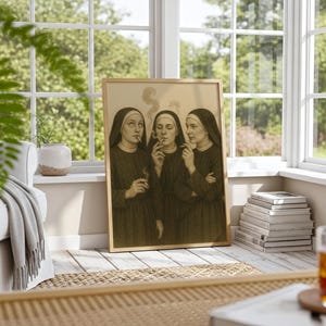 Puede incluir: Una impresi&oacute;n en tonos sepia enmarcada representa a tres monjas fumando cigarrillos. Las monjas visten h&aacute;bitos oscuros y tocas blancas. La obra de arte est&aacute; apoyada contra una pared en una habitaci&oacute;n soleada con una ventana y un sof&aacute;.