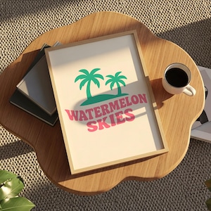 May include: A framed print with the words "Watermelon Skies" in pink, with green palm trees. The print is on a wooden table with a cup of coffee and some books.