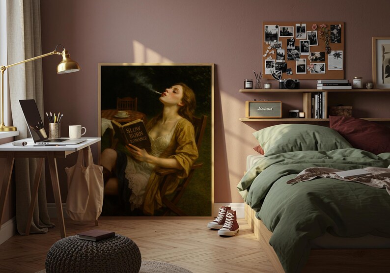 May include: A large framed artwork shows a woman reading a book titled "Slow Burn Romance" and smoking. The room includes a desk with a laptop, a bed with green bedding, and a corkboard displaying photographs.