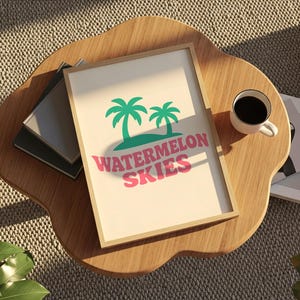 Op de afbeelding: Een ingelijste print met de woorden "Watermelon Skies" in roze, met groene palmbomen. De print ligt op een houten tafel met een kopje koffie en wat boeken.