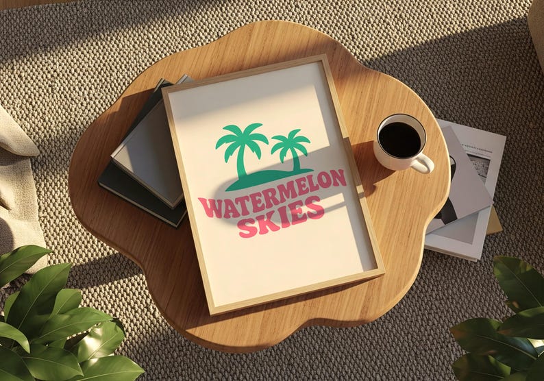 Op de afbeelding: Een ingelijste print met de woorden "Watermelon Skies" in roze, met groene palmbomen. De print ligt op een houten tafel met een kopje koffie en wat boeken.