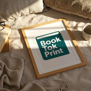 Może przedstawiać: Oprawiony wydruk z białym tekstem "Book Tok Print" na tle turkusowej ikony książki. Rama jest jasnobrązowa. Obok ramy znajdują się otwarta książka, okulary i ceramiczny kubek.
