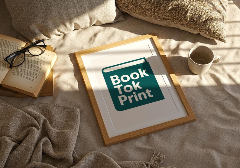 Może przedstawiać: Oprawiony wydruk z białym tekstem "Book Tok Print" na tle turkusowej ikony książki. Rama jest jasnobrązowa. Obok ramy znajdują się otwarta książka, okulary i ceramiczny kubek.